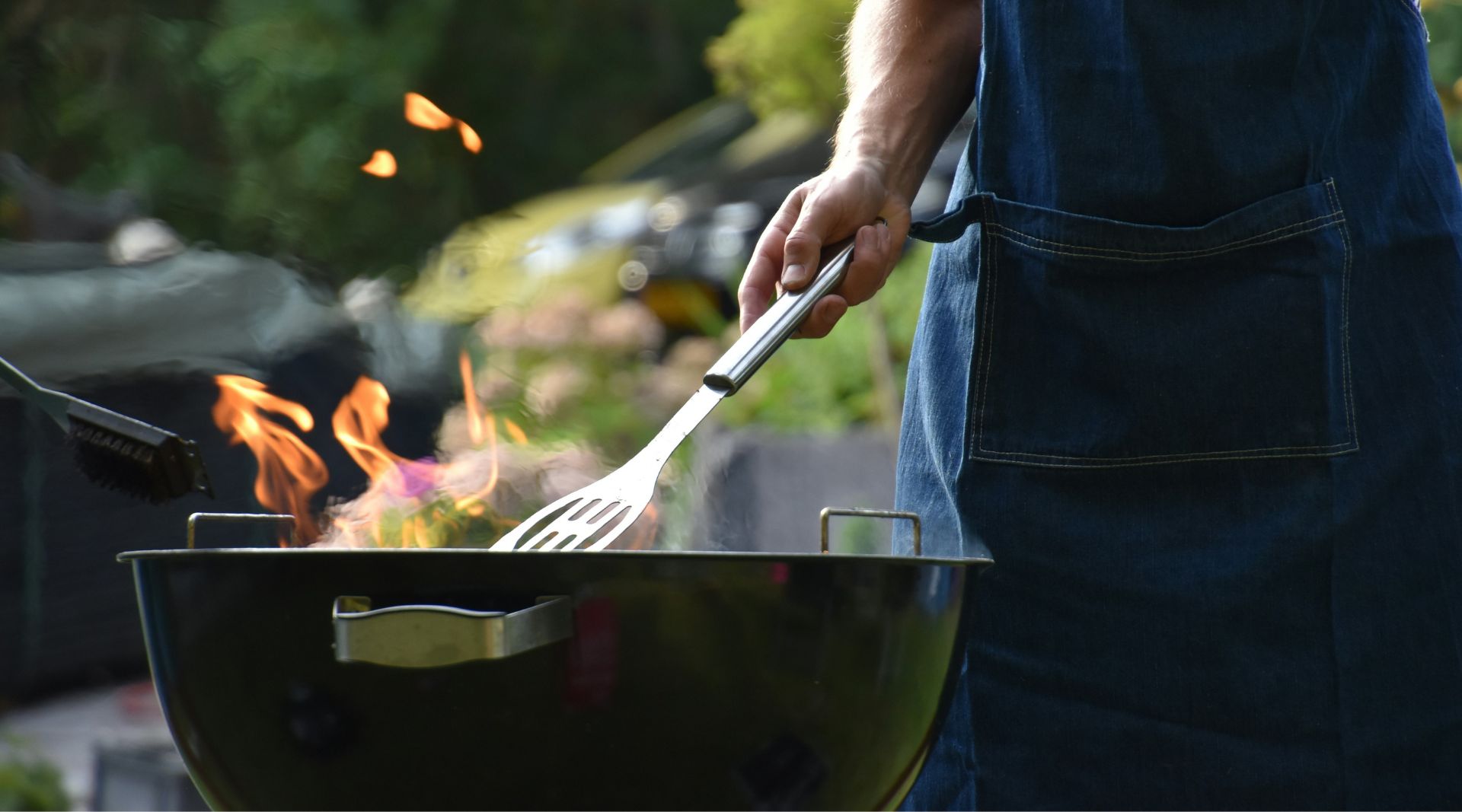 Person i forkle som griller med flammer på en kullgrill utendørs.