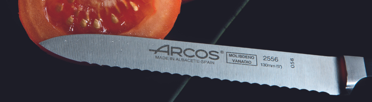 Nærbilde av skarpt ARCOS kjøkkenknivblad ved siden av skivet tomat, med fokus på knivens detaljer og merking, Made in Albacete Spain."